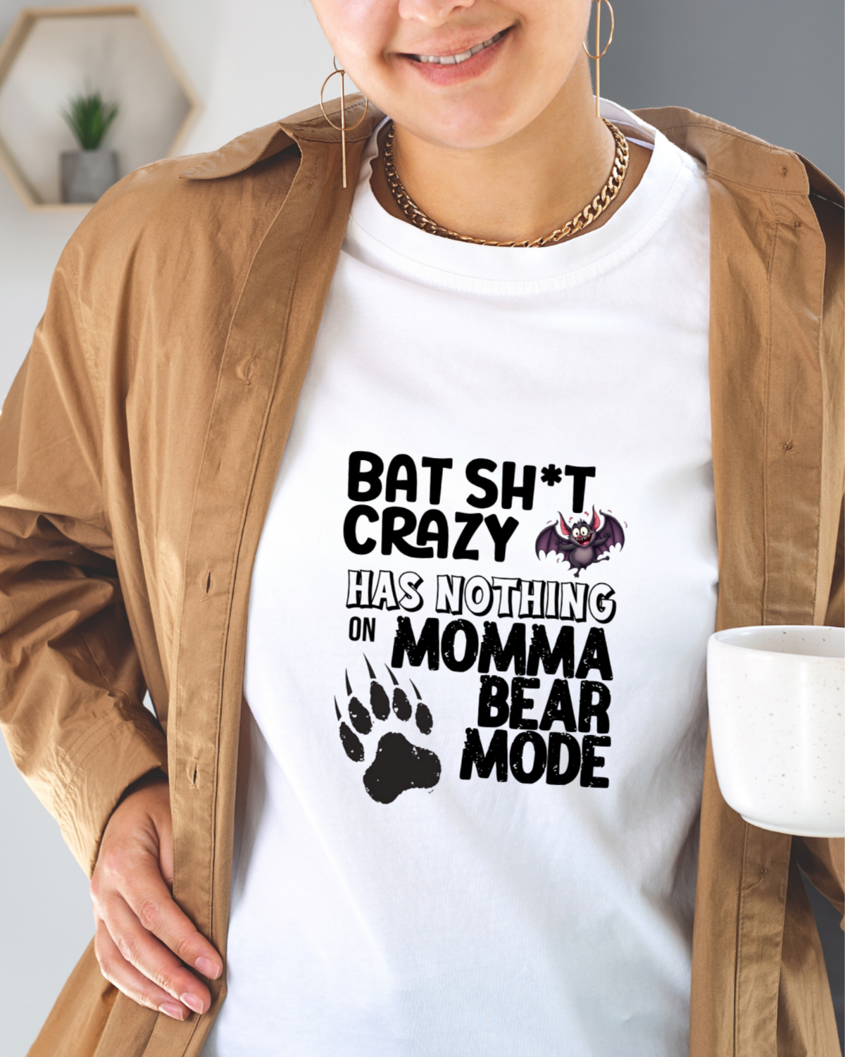 Person wearing a white t-shirt with a graphic and text reading "bat sh*t crazy has nothing on Momma bear mode" , holding a mug.