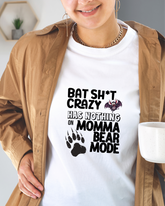 Person wearing a white t-shirt with a graphic and text reading "bat sh*t crazy has nothing on Momma bear mode" , holding a mug.